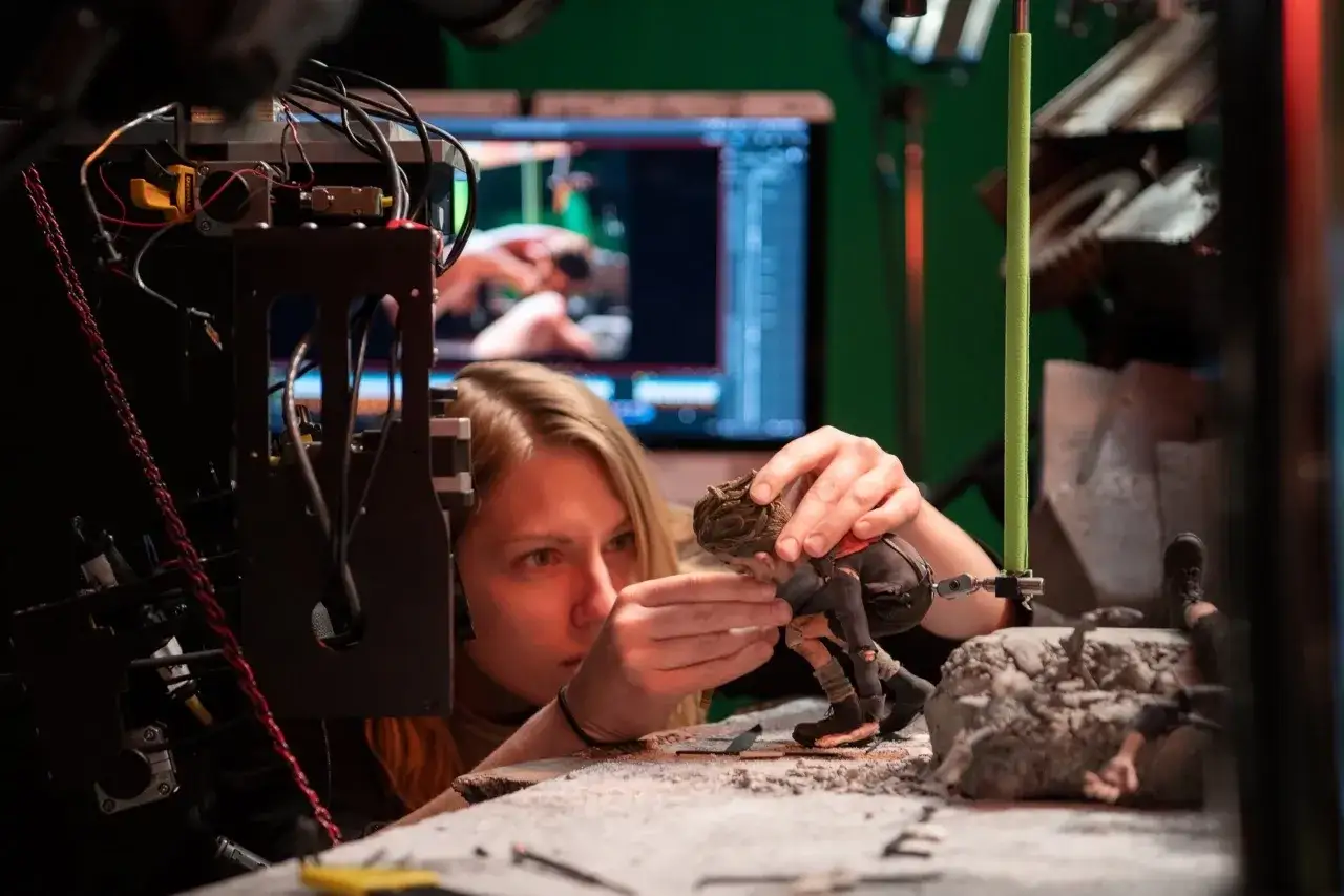 Tworzenie animacji poklatkowej: artystka precyzyjnie ustawia figurkę na planie filmowym, w tle monitor z podglądem sceny.