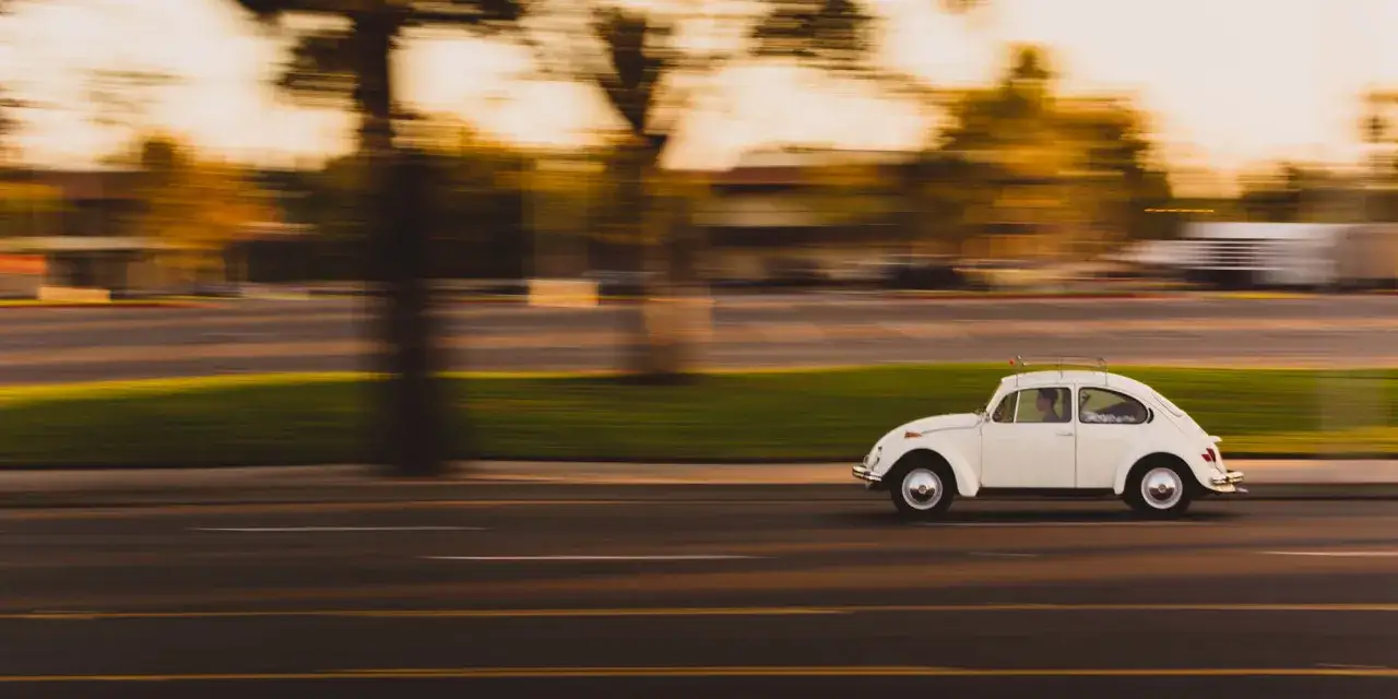 Biały Volkswagen Garbus w ruchu, uchwycony techniką panning. W tle rozmyte drzewa i budynki.